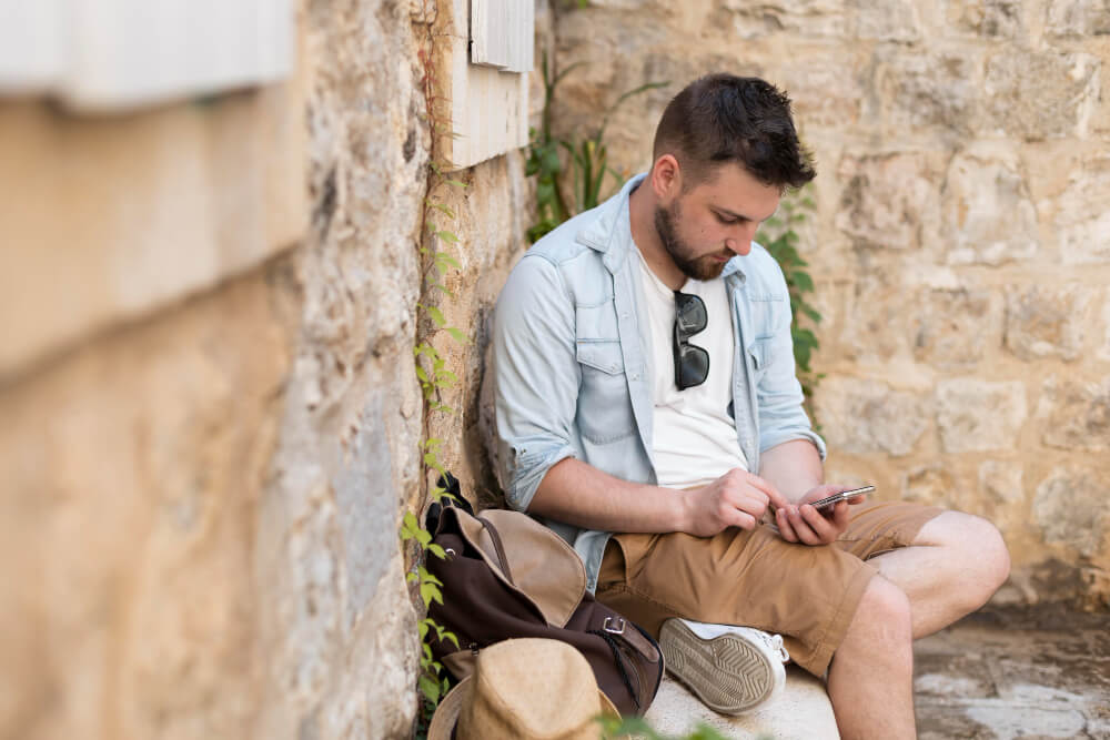 Mann mit kurzer Hose und Sonnenbrille sitzt an einer Hauswand